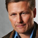 Photo of bestselling author David Baldacci with brown hair and a tight smile with a dark coat and brown scarf